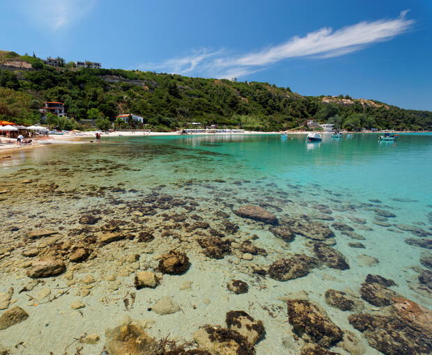 Clear turquoise water surrounds rocks in a tranquil bay with anchored boats. Sandy beach and lush green hills lie in the background, while people stroll along the shore under a blue sky.